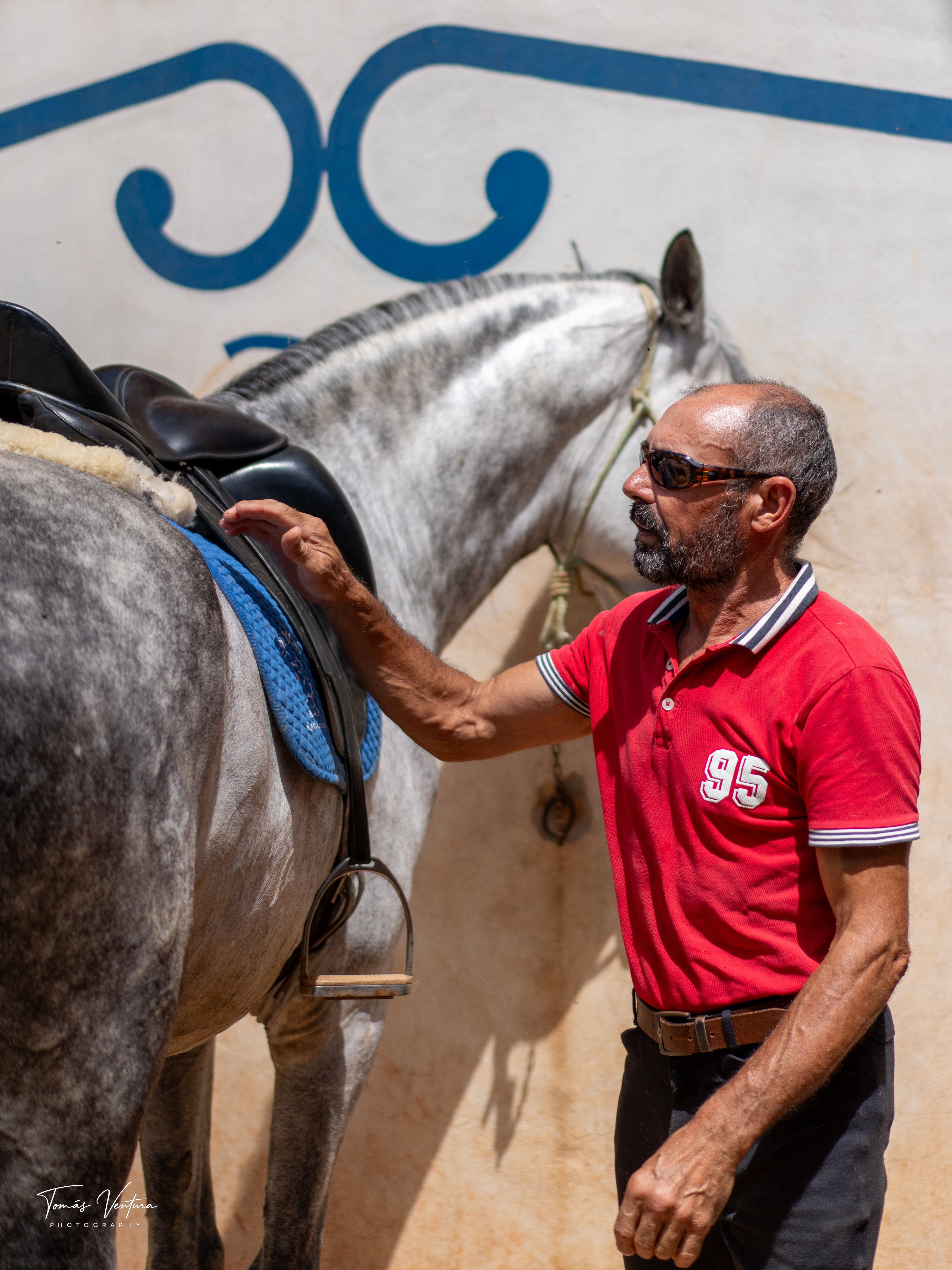 Jorge Ventura checks Oikos's saddle beside a white-and-blue wall.
